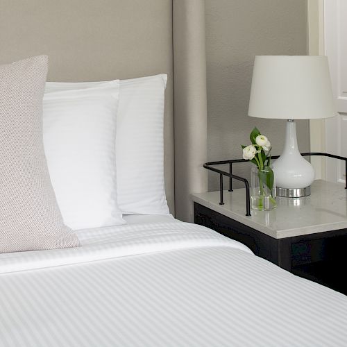 A neatly made bed with white linens and a beige pillow, next to a nightstand with a lamp and a small vase of white flowers on top.