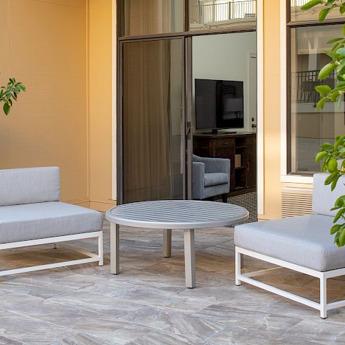 The image shows outdoor seating with two cushioned chairs and a round table on a patio, surrounded by leafy plants near a building with sliding glass doors.