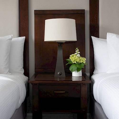 A neatly made hotel room with two white beds, a dark wooden nightstand in the middle, a lamp, and a small potted plant with white flowers on it.