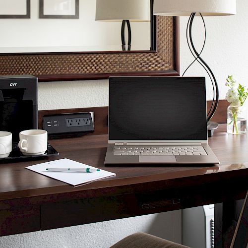 The image shows a desk with a laptop, a coffee machine, two cups, a notepad with a pen, and a lamp. There is a mirror and a small plant.