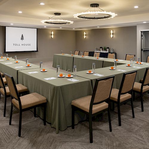 A conference room with U-shaped tables set with notepads, pens, and drinks, and a screen displaying 