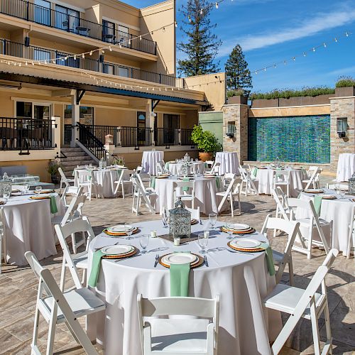 An outdoor dining area is set up with round tables, white chairs, and tablecloths, arranged for an event. There is a building in the background.