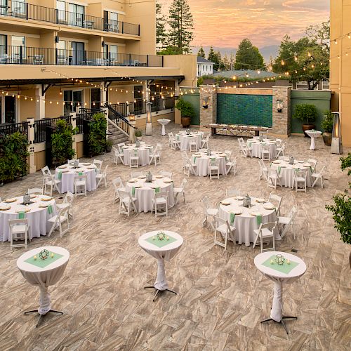 An outdoor event space with round tables and high cocktail tables, set with white tablecloths and chairs. String lights hang overhead.