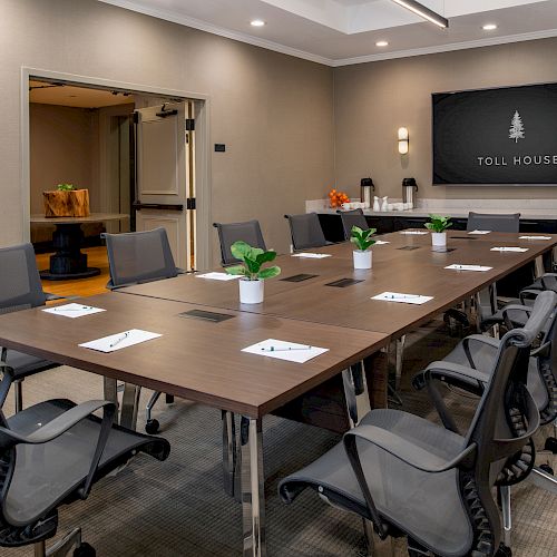 A conference room features a long table with chairs, notepads, potted plants, and a wall-mounted screen displaying 