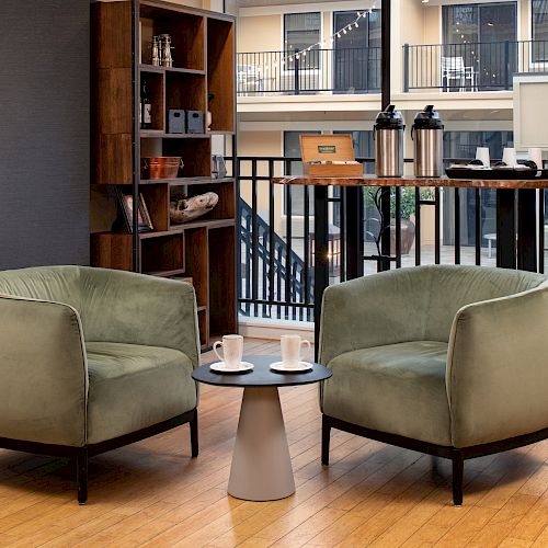 The image shows a cozy seating area with two green chairs, a small round table with two cups, and a wooden shelf and counter with coffee dispensers.