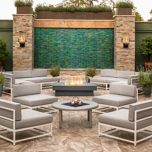 An outdoor seating area with modern gray furniture, a central fire pit, greenery in the background, and a textured blue wall feature ending the sentence.