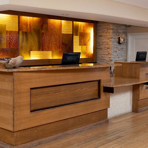 The image shows a modern hotel reception desk with wood accents, warm lighting, and two computer monitors on the counter.