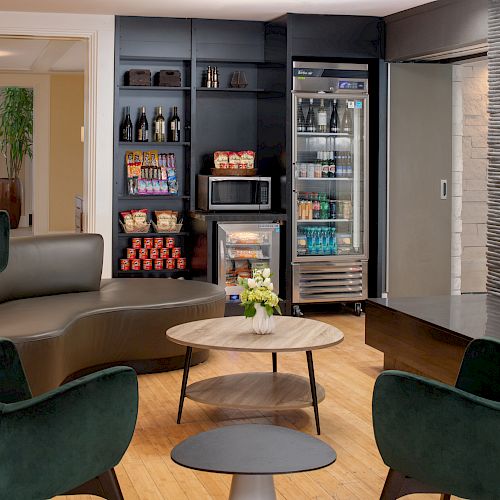 A modern lounge area with black shelves stocked with snacks and drinks, a fridge, microwave, green chairs, and a wooden coffee table with flowers.