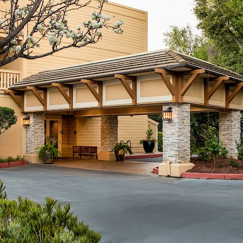 The image shows the entrance of a building with a covered walkway, stone columns, surrounding trees and plants, and a well-maintained driveway.